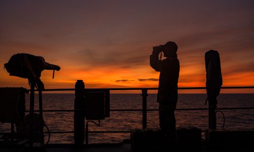 Silhouette of a uniformed naval personnel looking through binoculars on a ship at sea during a vibrant orange and purple sunset.