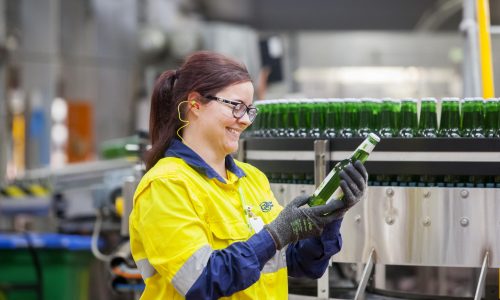 A smiling factory worker in high-visibility gear inspects a green glass bottle on a production line, highlighting quality control and safety in a modern manufacturing facility.